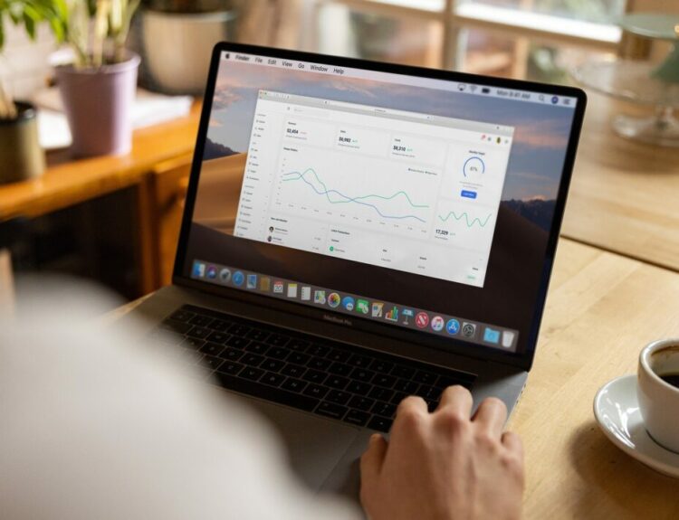 Osoba pracująca na laptopie z otwartym panelem CRM, ukazującym analizę danych, wskaźniki wydajności oraz statystyki sprzedaży. W tle znajduje się biurko z filiżanką kawy oraz rośliny doniczkowe.
