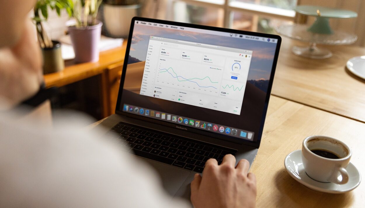 Osoba pracująca na laptopie z otwartym panelem CRM, ukazującym analizę danych, wskaźniki wydajności oraz statystyki sprzedaży. W tle znajduje się biurko z filiżanką kawy oraz rośliny doniczkowe.